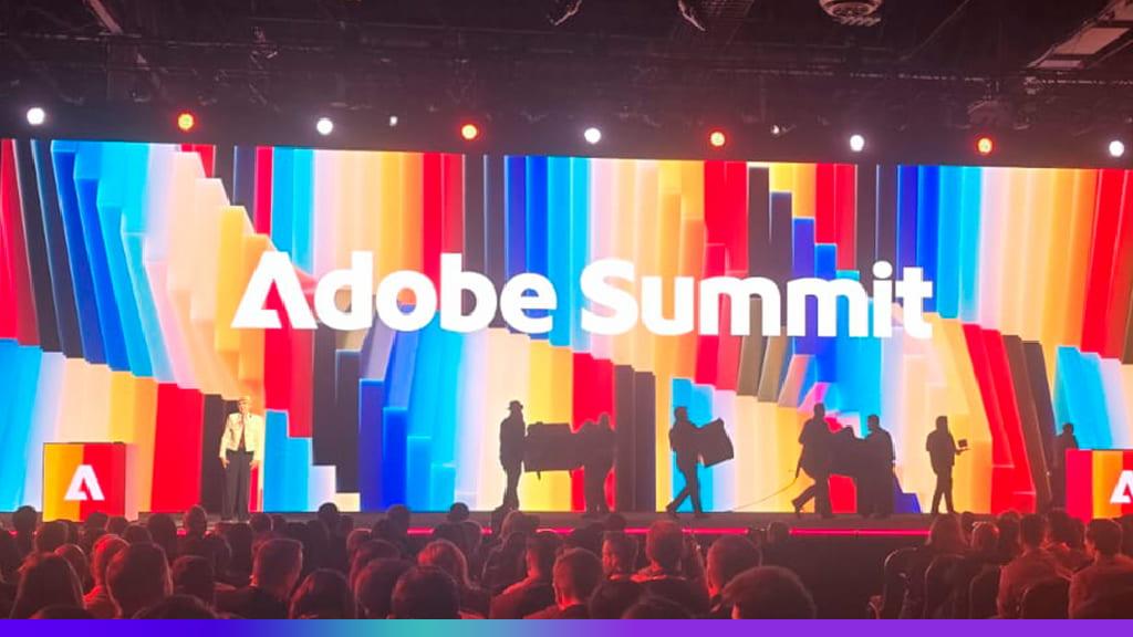 Painel de palco iluminado com o logo do Adobe Summit em destaque, com uma tela de fundo colorida e várias pessoas no palco, em um evento de tecnologia.